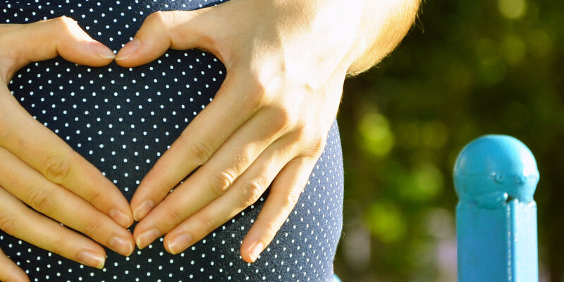 Hände formen ein Herz auf dem Bauch einer schwangeren Frau.